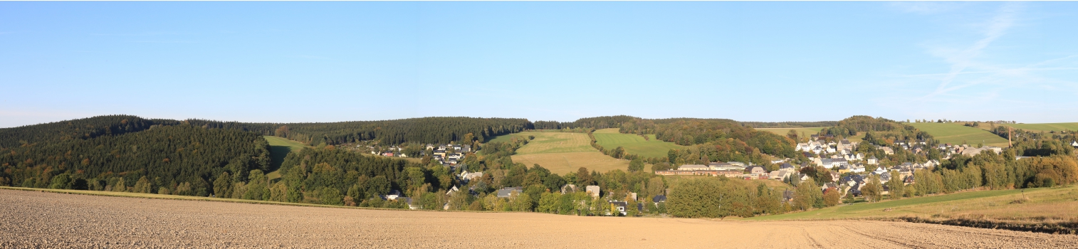Das Foto zeigt eine weitläufige, ländliche Landschaft mit landwirtschaftlichen Feldern und Wäldern. In der Mitte der Ort Auerbach, der sich malerisch in das Tal schmiegt. Die Farben deuten auf den Herbst hin - Warme Farben, leicht gedämpftes Licht und eine ruhige Atmosphäre. Die Perspektive ist panoramisch, wodurch man ein Gefühl für die Weite und Ruhe der Region bekommt - fast so als würde man von einem Aussichtspunkt auf das Tal hinabblicken.